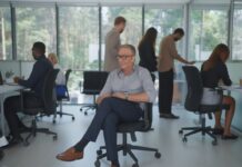 Simposio de EE.RR. para la sensibilización de los medios de comunicación Un hombre de mediana edad, sentado en una silla con ruedas, en el centro de una oficina con grandes ventanales y con otros colaboradores
