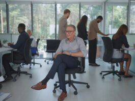 Un hombre de mediana edad, sentado en una silla con ruedas, en el centro de una oficina con grandes ventanales y con otros colaboradores