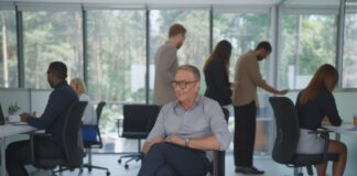 Un hombre de mediana edad, sentado en una silla con ruedas, en el centro de una oficina con grandes ventanales y con otros colaboradores